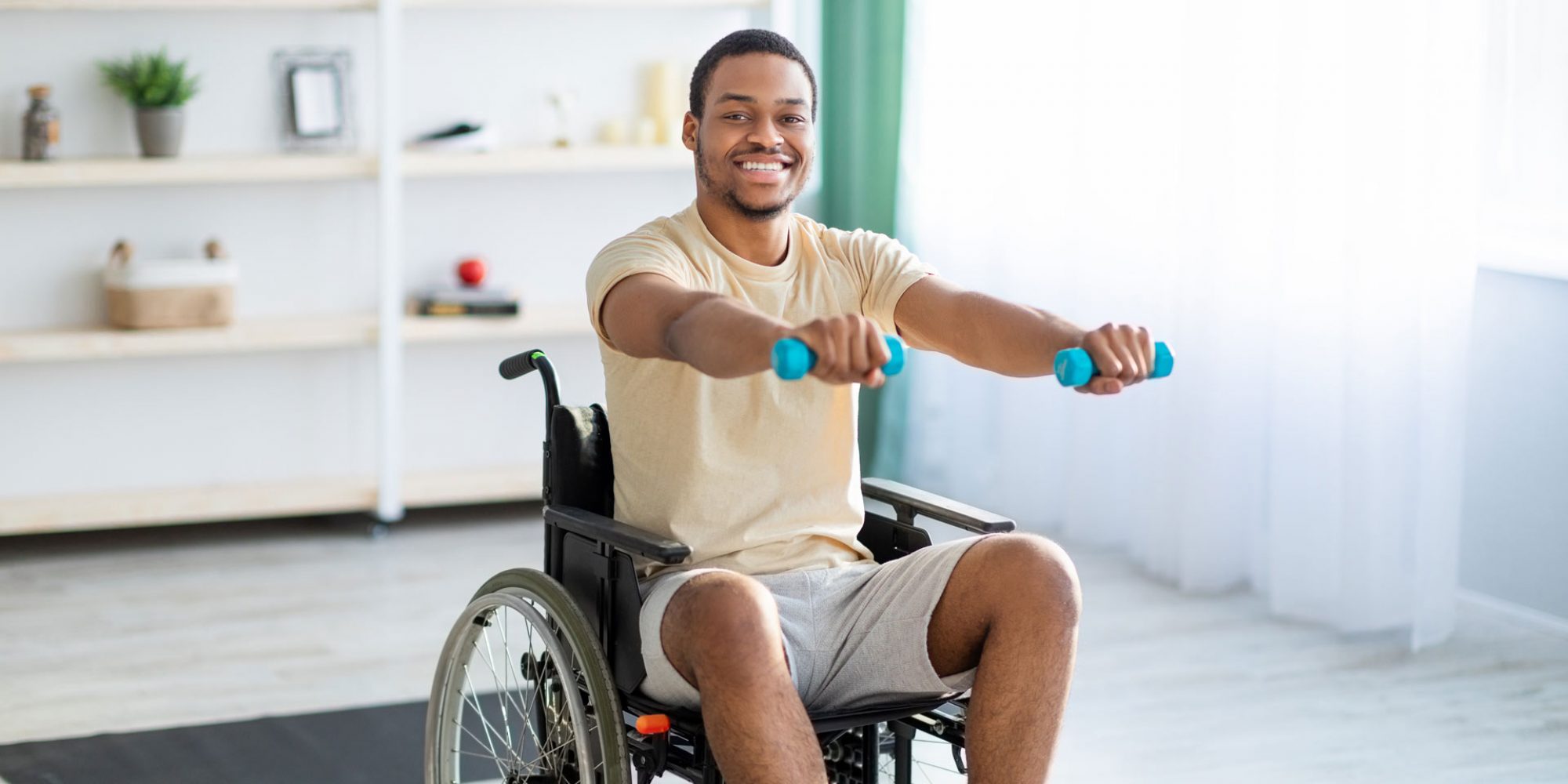 Mann sitzt lachend im Rollstuhl und macht Muskelübungen mit kleinen Gewichten