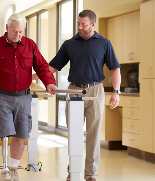 Mann läuft mit einer Beinprothese mit Hilfe von einem zweiten Mann an einem Geländer entlang